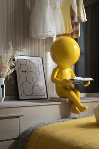 A modern children's bedroom featuring a yellow decorative figure reading a book, with a framed art piece and dried plants on