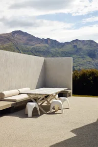 An outdoor lounge area with concrete walls, a wooden table, and white stools, set against a mountainous backdrop.