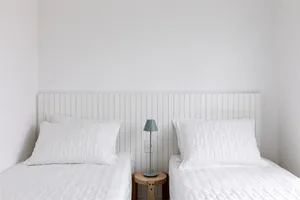 A minimalist bedroom with two beds, white bedding, and a small wooden table with a lamp.