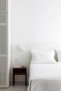 A minimalist bedroom featuring a white quilted blanket on a bed with a white headboard and wooden nightstand against a white 