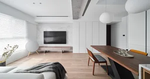 A minimalist open-plan living room with a large TV mounted on a white marble wall, features a wooden dining table, and clean,