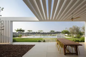A modern outdoor space with concrete and stone elements, featuring a wooden table and chairs, a swimming pool, and lush green