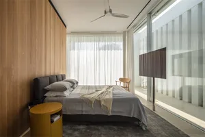 A modern minimalist bedroom featuring a wooden wall and floor, large glass windows, and a comfortable bed with grey bedding a