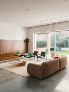 A modern living room with concrete flooring, a brown sectional sofa, a round glass coffee table, and a wooden shelf with deco
