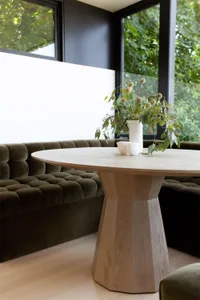 A modern living room featuring a wooden table adorned with greenery and two white cups, situated in a corner with large windo