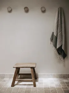 A minimalist indoor wooden stool placed against a tiled floor and plain wall, with a grey and green towel draped on the wall.