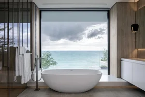 A modern bathroom with a large window overlooking the ocean, featuring glass tiles and a freestanding bathtub.