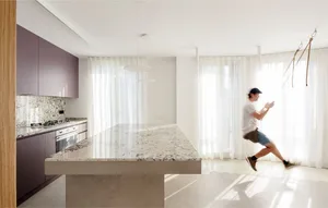 A modern kitchen features a large granite marble countertop and island with purple cabinets and white curtains in the backgro
