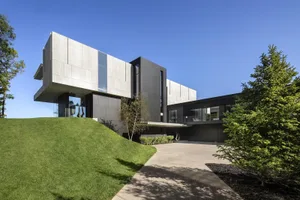 Modern, minimalist house with concrete and glass exterior, set on a grassy hillside.