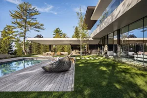A modern residential exterior featuring concrete and wooden structures, surrounded by greenery and a swimming pool.