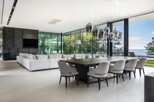 A modern living and dining room with white furniture, wooden table, and large windows overlooking the sea.