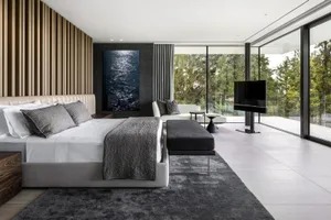 A modern bedroom featuring dark textiles, a glass TV, and a view of nature.
