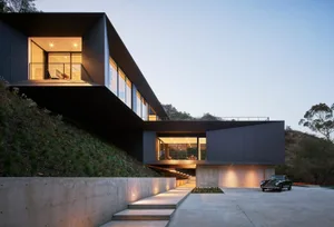 Modern metal-clad cabin with garage and warm interior lights, set against a hillside.