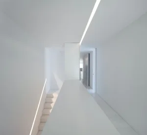 A minimalist white interior with a white wall and floor, featuring a staircase with white steps and a light source from above