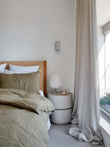 Modern minimalist bedroom featuring a bed with neutral bedding and a round bedside table with a lamp and alarm clock, alongsi