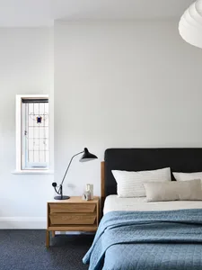 Modern bedroom featuring a wooden nightstand with a lamp, a bed with a fabric headboard and neutral bedding, and a small wind