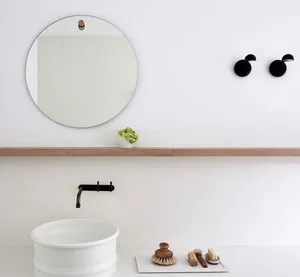 A minimalist bathroom featuring a white ceramic sink, a round mirror with a wooden holder, and a shelf with black hooks and a