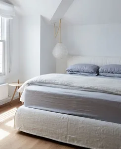 A minimalist bedroom with a bed dressed in light blue linen blankets and pillows, featuring a pendant light and a window with