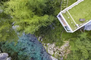 A modern hillside home featuring wood, glass, and stone materials surrounded by trees and a river.