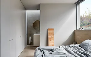 Minimalist bedroom with concrete walls and floor, featuring a bed and a small sink with a circular mirror.