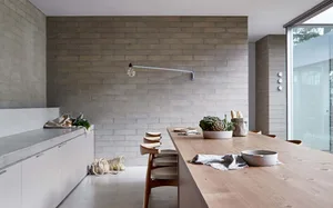 A modern minimalist kitchen with a wooden dining table and concrete countertops, featuring a brick wall and natural light.