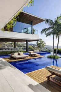 A modern villa featuring a large outdoor pool with lounge chairs and a glass and wood patio overlooking a scenic landscape.