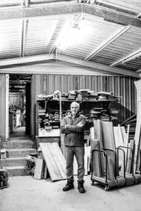 An older man wearing a gray fleece jacket and jeans, standing in a workshop surrounded by wood and metal