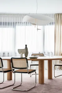 A modern dining room with a wooden table, black and rattan chairs, and a large window with sheer curtains.