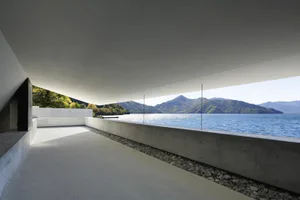 現代簡約室內空間，混凝土地板與牆面，眺望寧靜水岸景觀，遠處有山巒