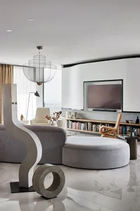 A modern living room with concrete, wood, and fabric materials, featuring a round ottoman, a bookshelf, and a television.