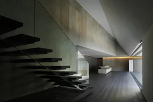 Modern minimalist residential interior with concrete walls and floor, black floating stairs, and a kitchen area.