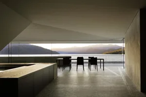 Modern residential interior with concrete walls and floor, featuring a dining table and chairs overlooking a lake and mountai