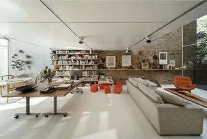 A minimalist living room with a concrete floor, a long sofa, red stools, and a bookshelf.