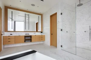 A modern bathroom with white marble walls, countertops, and floor tiles, featuring a large mirror, wooden vanity, and a glass