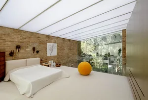 A minimalist bedroom with a white bed, yellow ball, and glass windows showing a garden.