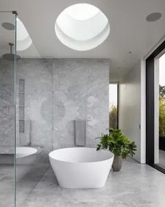 Modern bathroom interior with a freestanding white bathtub, marble walls, and a large skylight.