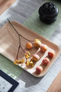 A wooden tray with small, round, yellow and pink fruits, a yellow flower stem, and a ceramic incense burner on a green and gr
