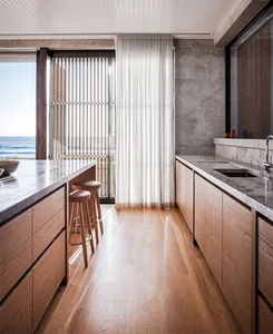 Modern kitchen with wooden cabinets, marble countertops, and a view of the ocean through large windows with vertical blinds a