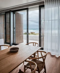 A minimalistic dining room with a wooden table, chairs, and a view of the ocean through large glass doors.