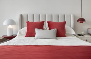 A minimalist bedroom with a white upholstered bed, red and white accent pillows, and a white bedspread.