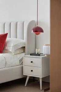 A minimalist bedroom with a white leather bed and a white bedside table with a red pendant light hanging above it.