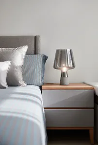 A modern bedroom featuring a wooden headboard, concrete bedside table, and glass lamp.
