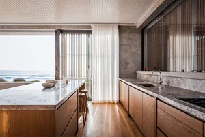 A modern residential kitchen featuring marble countertops and wooden cabinets with a view of the ocean through large windows 