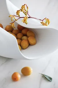 A modern ceramic bowl filled with fresh apricots and adorned with yellow flowers, set on a white marble surface.