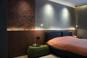A minimalist bedroom featuring a brick wall, a black bed frame, and a green ottoman.