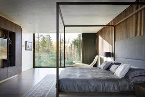 Modern bedroom with a canopy bed made of wood, featuring grey textiles and a view of the forest through large windows.