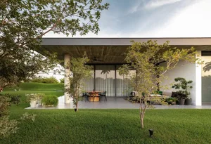 A modern house with a concrete exterior has a large open-air patio with a dining table and chairs surrounded by greenery and 