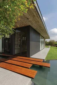 Modern residential pool with wooden deck and concrete roof structure, surrounded by greenery.