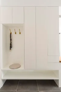 A minimalist wardrobe closet with white cabinets, brass hooks, and a cushion seat.