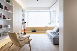 A minimalist living room with wooden flooring and furniture, featuring a low-profile sofa, a woven armchair, and a built-in b